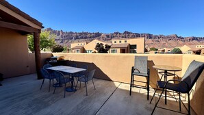 A patio with a table and chairs, a grill, and a view of a residential area with houses and mountains in the background. - Coyote Run @ Rim Village - Pet Friendly (Moab)