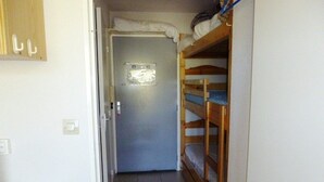 Een smalle gang met een deur aan het einde, een houten stapelbed aan de rechterkant en een klein houten kastje aan de linkerkant.