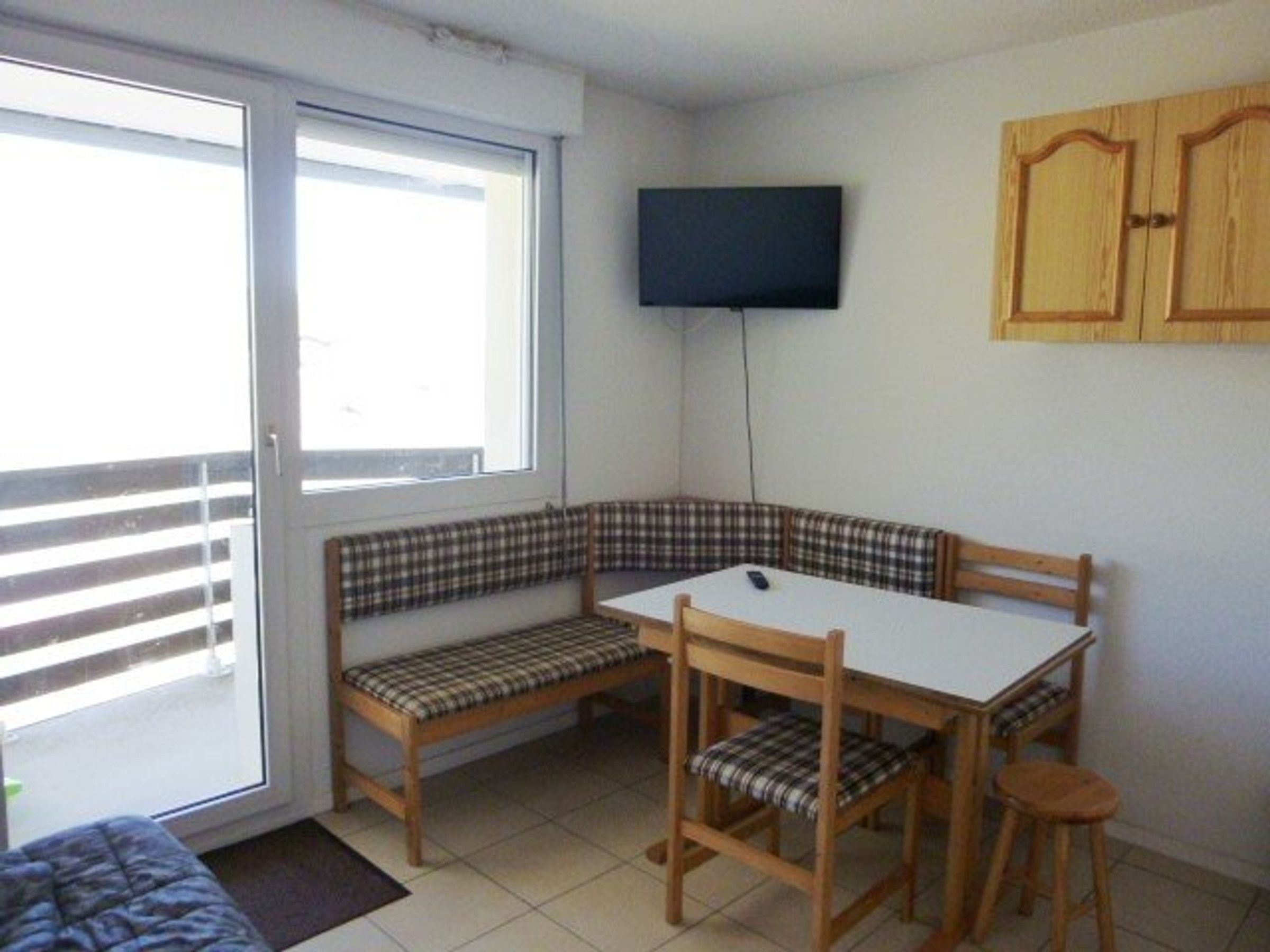 Una habitación pequeña con una banca, una mesa, una silla, un televisor montado en la pared y un ventanal corredizo.