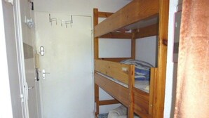 Een kleine, eenvoudige kamer met een stapelbed, een deur met hendel en een witte muur.