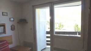 Een kamer met een rode stoel, een klein tafeltje, een boekenkast, een klok en een schuifdeur die uitkomt op een buitenruimte met uitzicht op bomen en een hek.