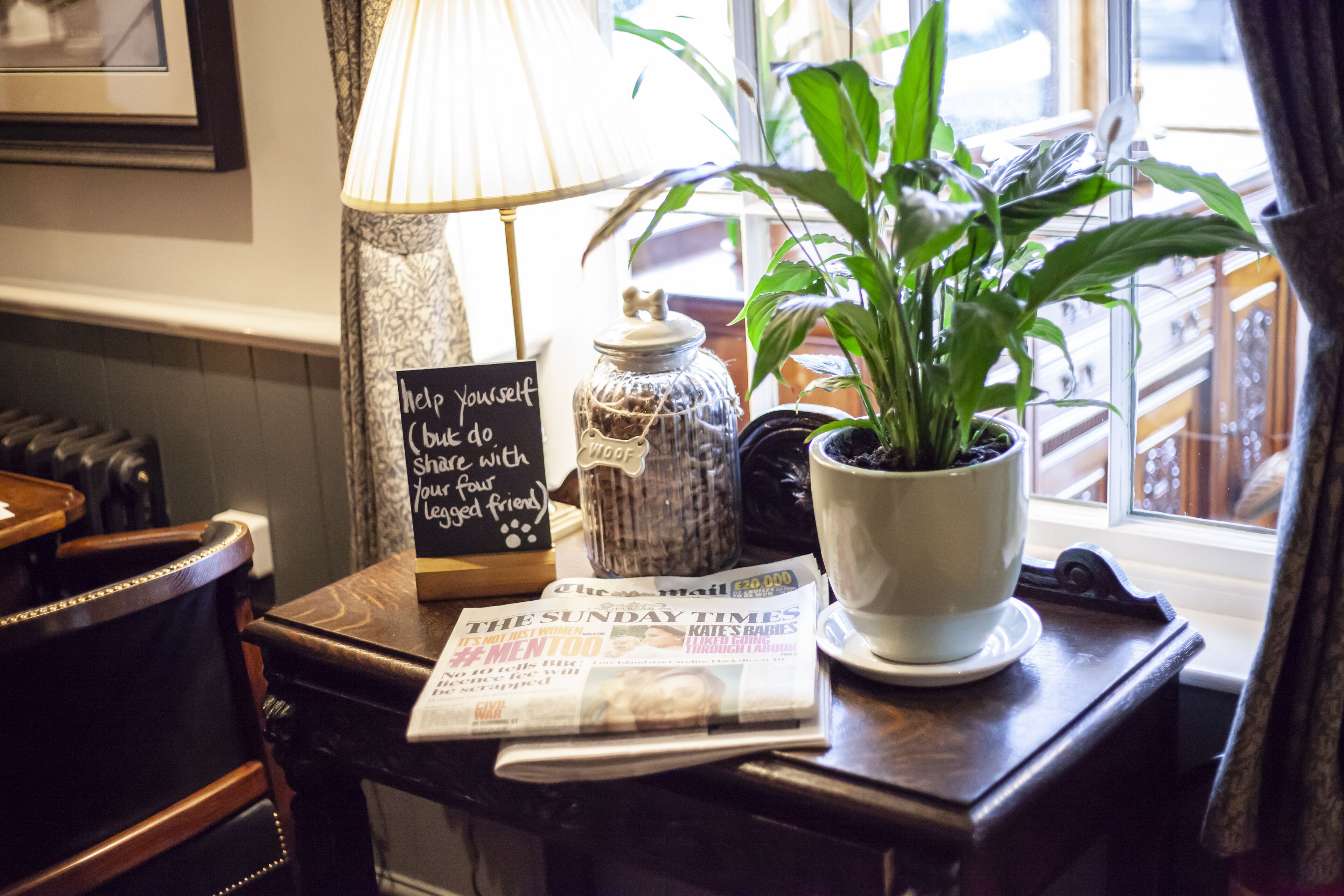 Una mesa con un periódico, una lámpara, una planta en maceta y un frasco decorativo.