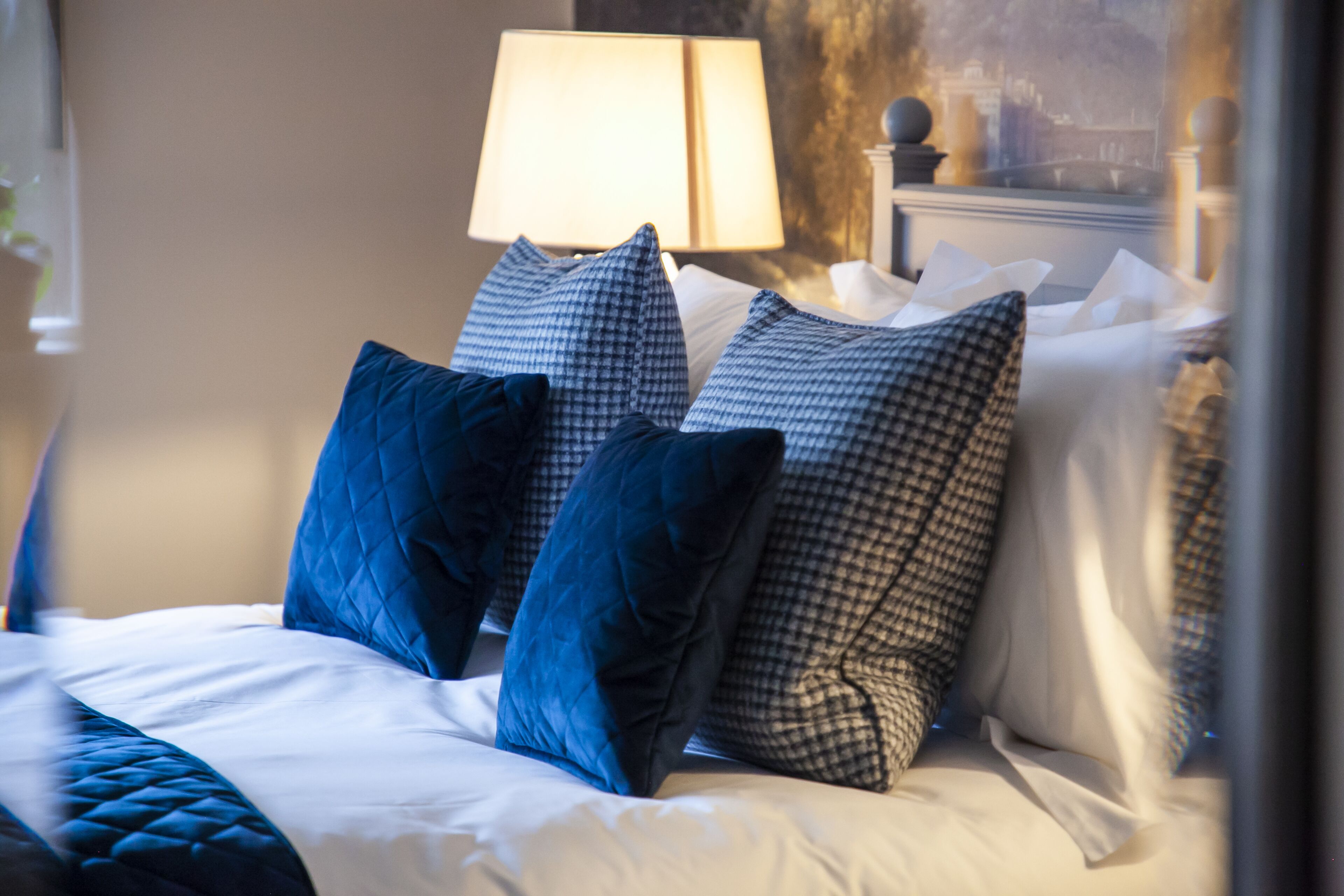 Ein ordentlich bezogenes Bett mit blauen und karierten Kissen, einer weißen Tagesdecke und einer Lampe im Hintergrund.