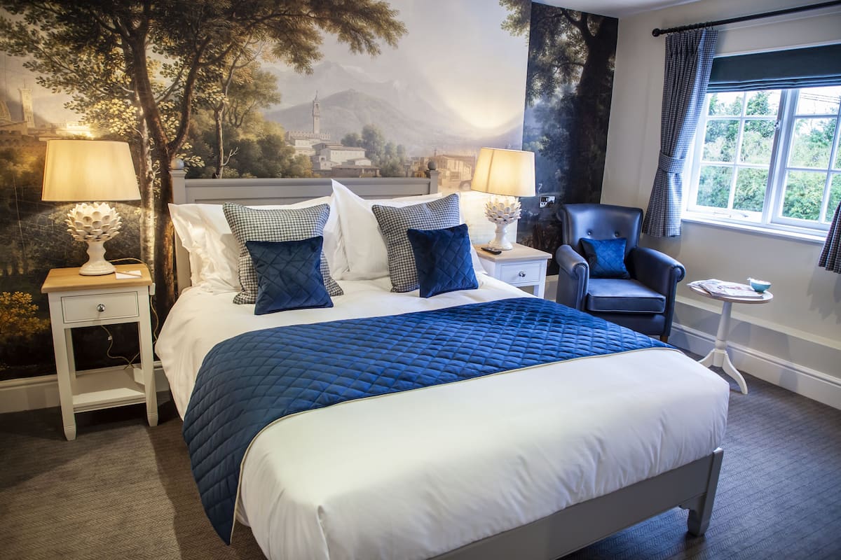 Un lit bien fait, recouvert d’une couette bleue, deux tables de chevet avec des lampes, une chaise et une fenêtre avec des rideaux.