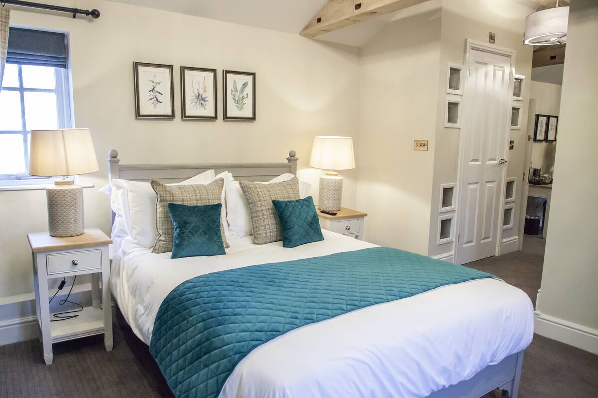 Un lit bien fait, recouvert d’une courtepointe turquoise, avec deux lampes de chevet et une fenêtre ornée d’un rideau.