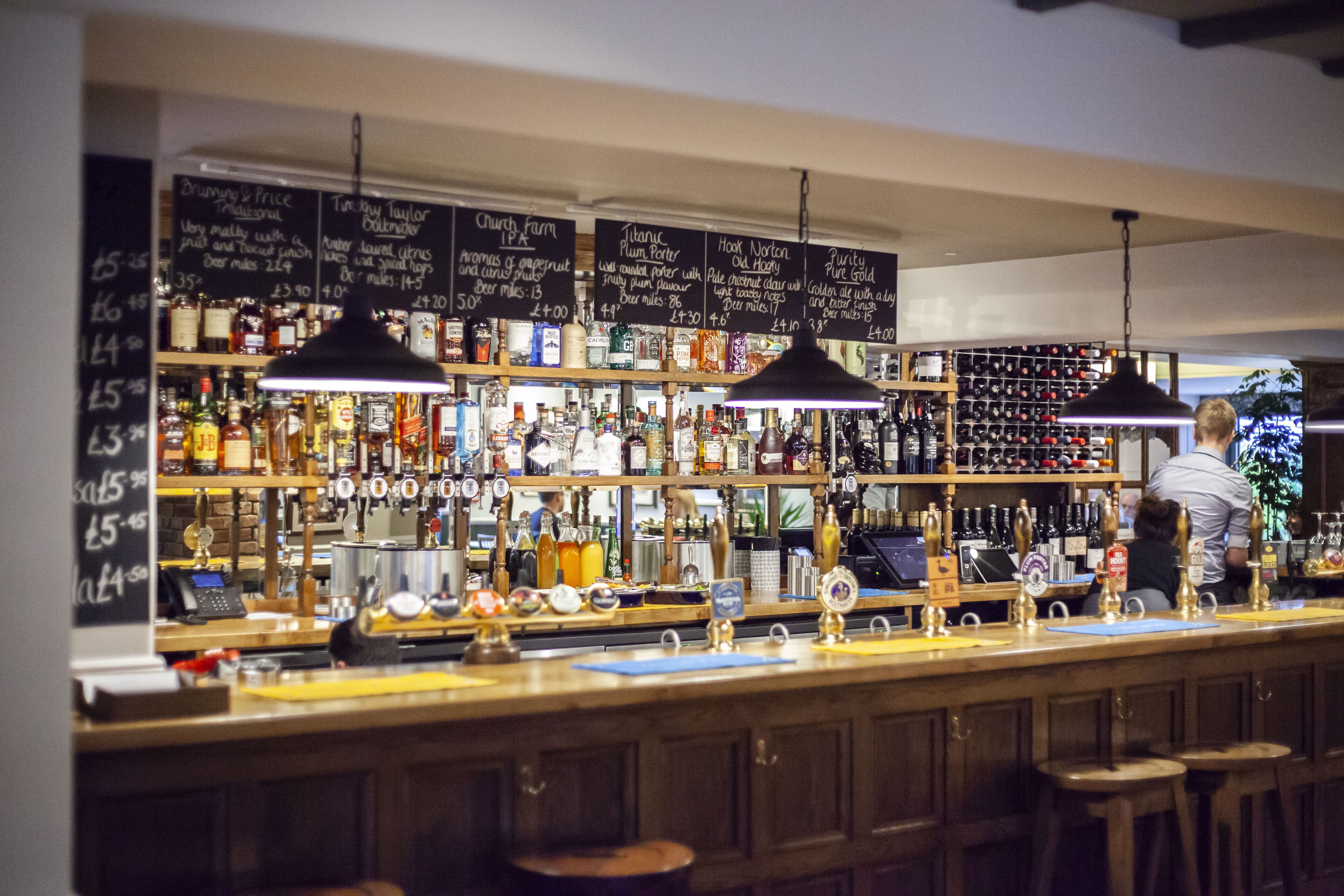Un bar con numerosi rubinetti, scaffali pieni di bottiglie e una lavagna con il menu delle bevande e i relativi prezzi.