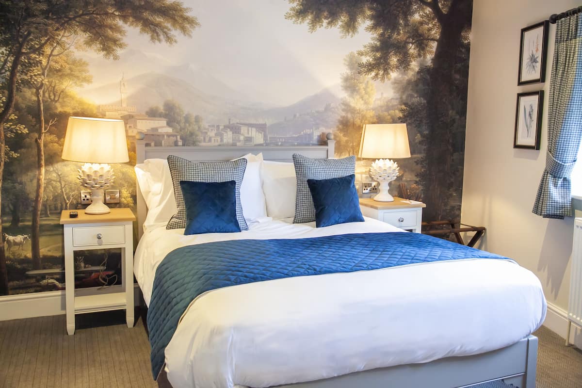 Une chambre à coucher avec un grand lit, deux tables de chevet avec des lampes, une fresque représentant un paysage et une fenêtre ornée d’un rideau à carreaux.