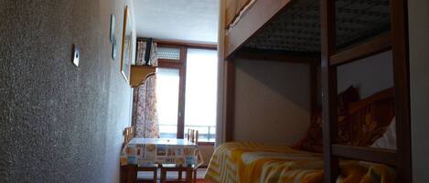 A compact room with a bunk bed, a dining table with chairs, and a window with curtains.