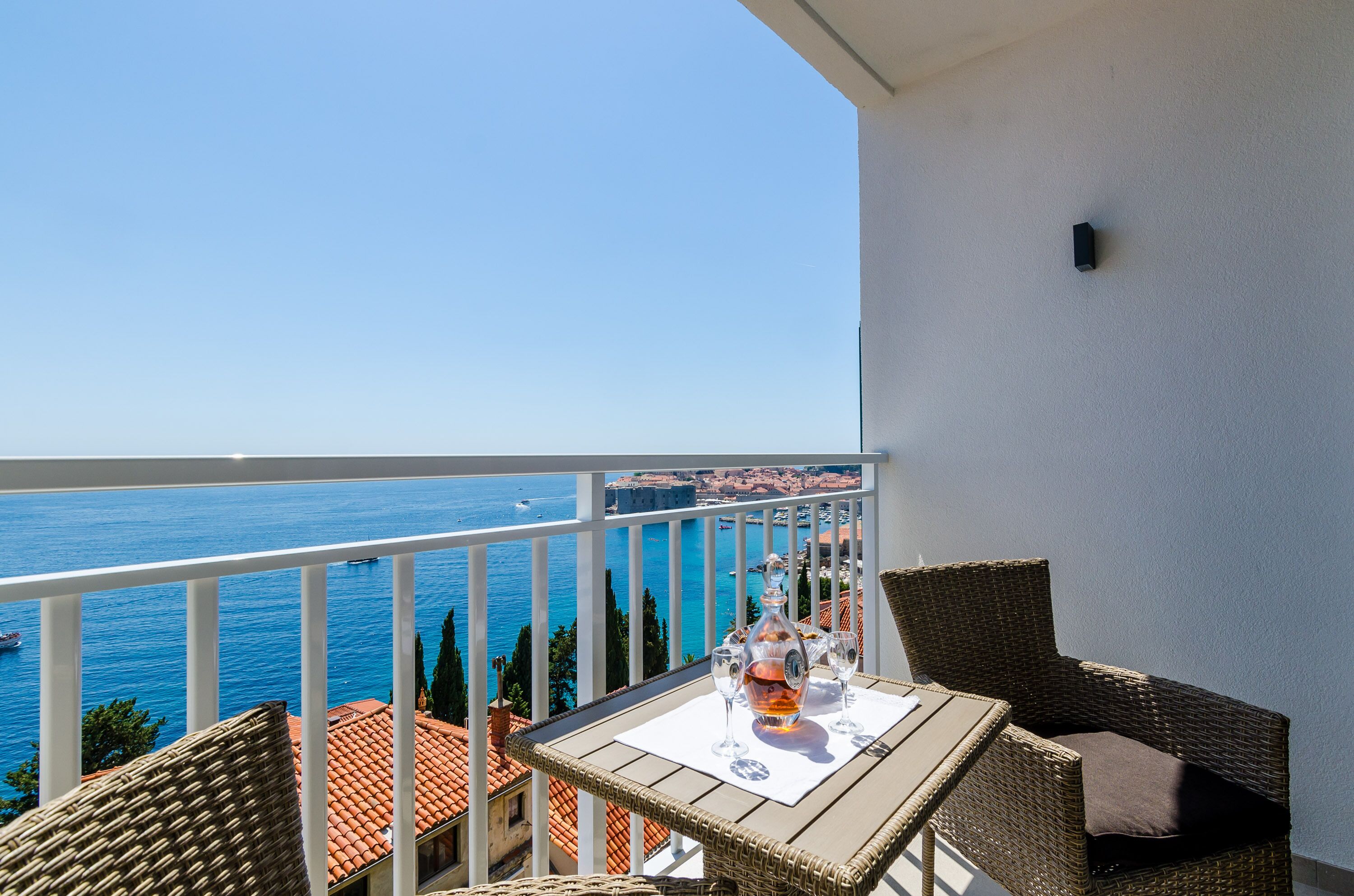 A balcony with wicker furniture, a table with a bottle and glasses, and a view of the sea.