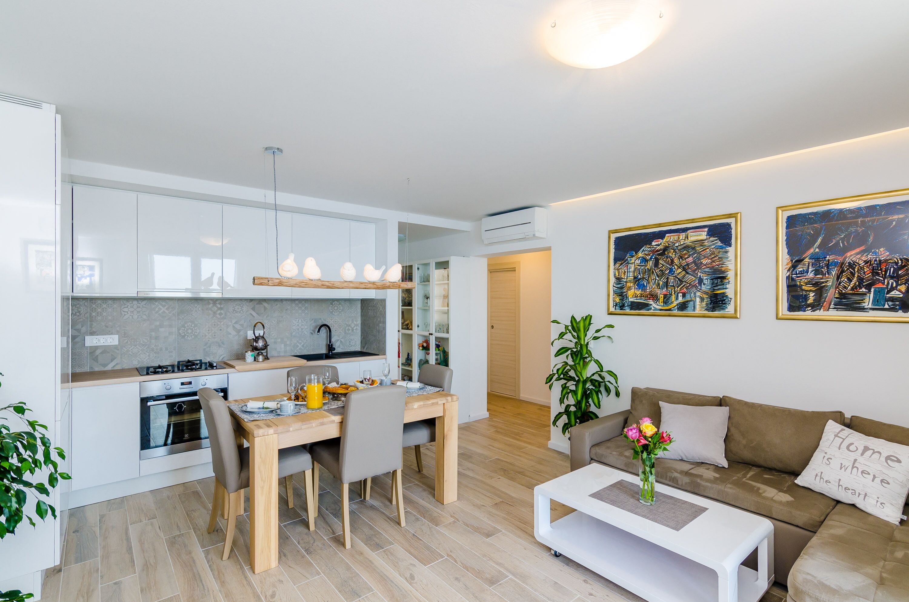A modern kitchen with a dining area, a living room with a sofa and a coffee table, and two framed artworks on the wall.