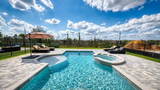 A swimming pool with two hot tubs, surrounded by lounge chairs and an umbrella, with a clear sky and trees in the background.