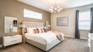 A bedroom with a tufted headboard bed, bedside tables, a large mirror, a chandelier, and a window with curtains.