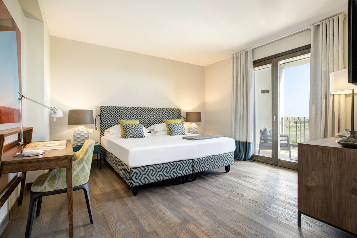 A hotel room with a large bed, a desk with a lamp, a chair, and a view of the outdoors through a sliding door.