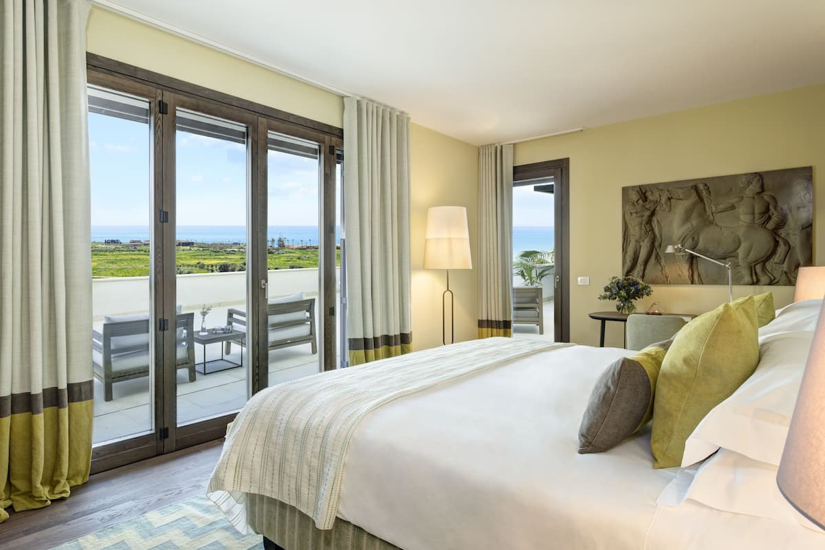 A hotel room with a bed, a desk, a chair, and a view of the sea through large sliding doors.