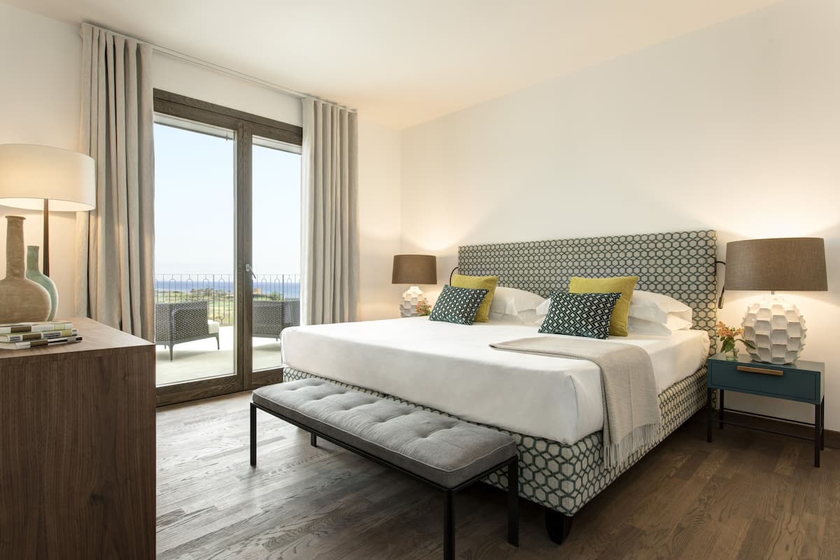 A hotel room with a large bed, a bench, two bedside tables with lamps, a desk, and a view of the outdoors through a sliding door.