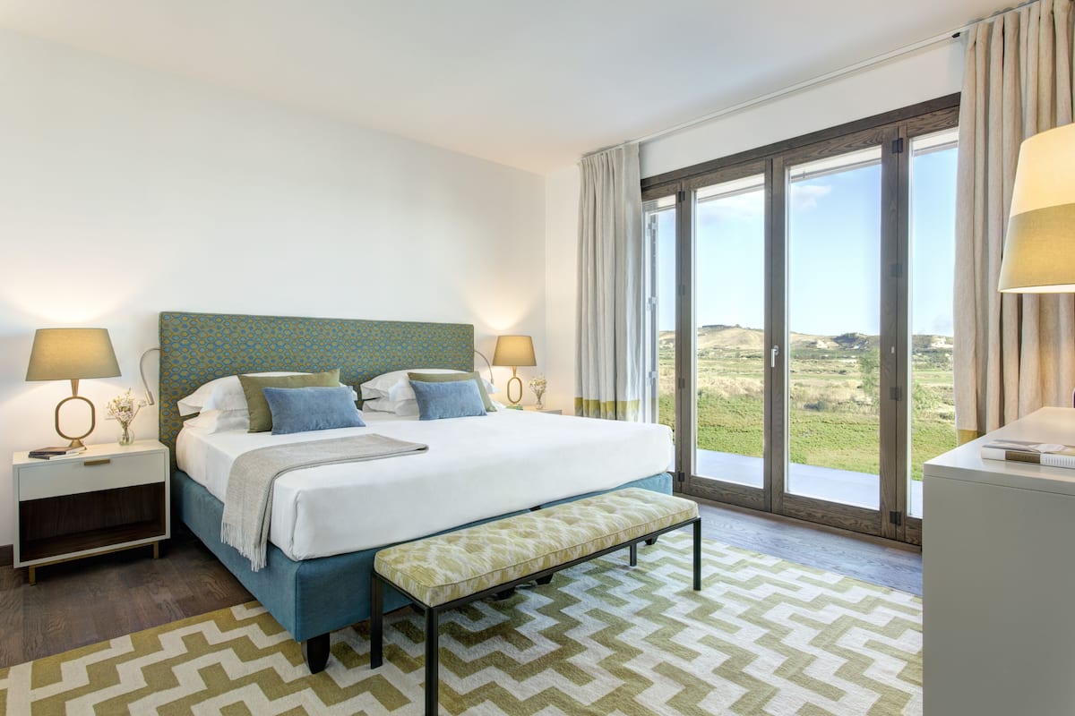 A bedroom with a large bed, two bedside tables, a bench, and a view of the outdoors through a sliding glass door.