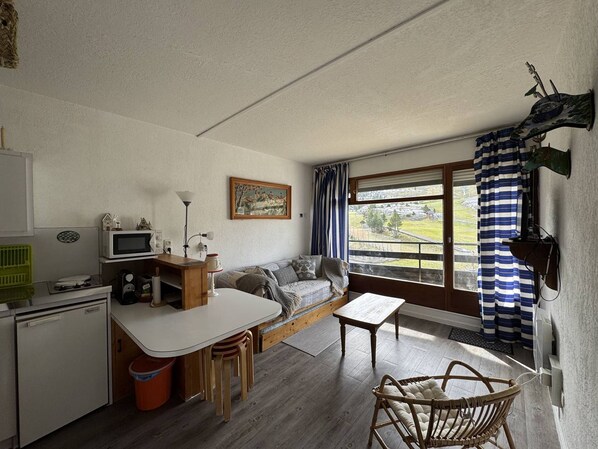 A compact living space with a kitchenette, a sofa, a dining table, and a view of a green landscape through a window.
