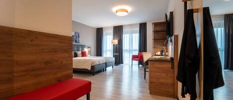 A modern hotel room with a wooden headboard, a red bench, a bed with a white duvet, a desk with a lamp, and a mirror with hanging clothes.