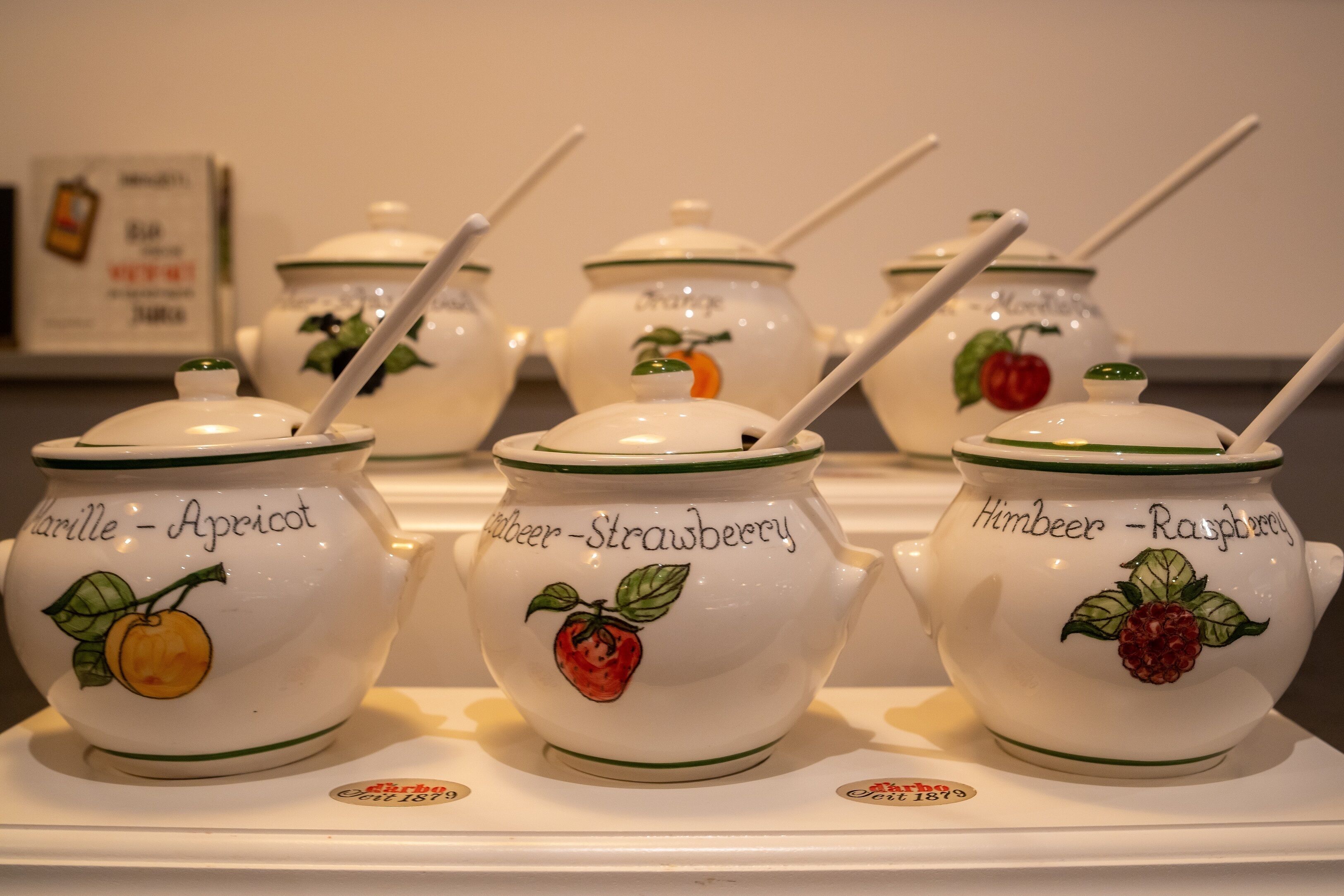 A display of white ceramic jars with green trim and fruit illustrations, each labeled with a different fruit name.