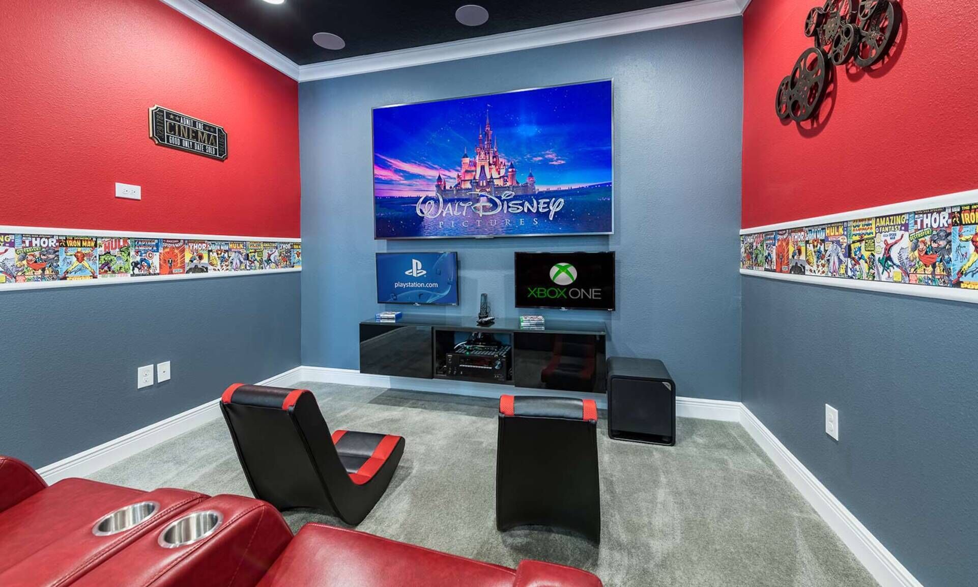 Een bioscoopzaal met een grote televisie waarop 'Walt Disney Pictures' staat, twee gameconsoles en rode en zwarte gamingstoelen.