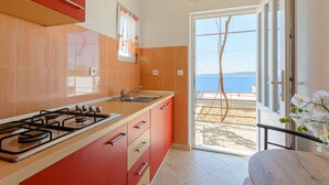 A modern kitchen with a gas stove, sink, and red cabinets. A sliding glass door leads to a balcony with a view of the sea. - Rooms Sani (Brela)