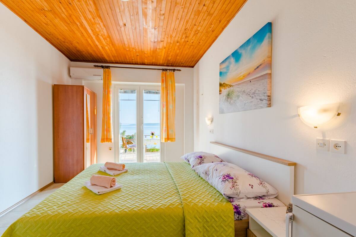 A bedroom with a wooden ceiling, a bed with a green quilt, a painting of a beach, and a balcony with a view.