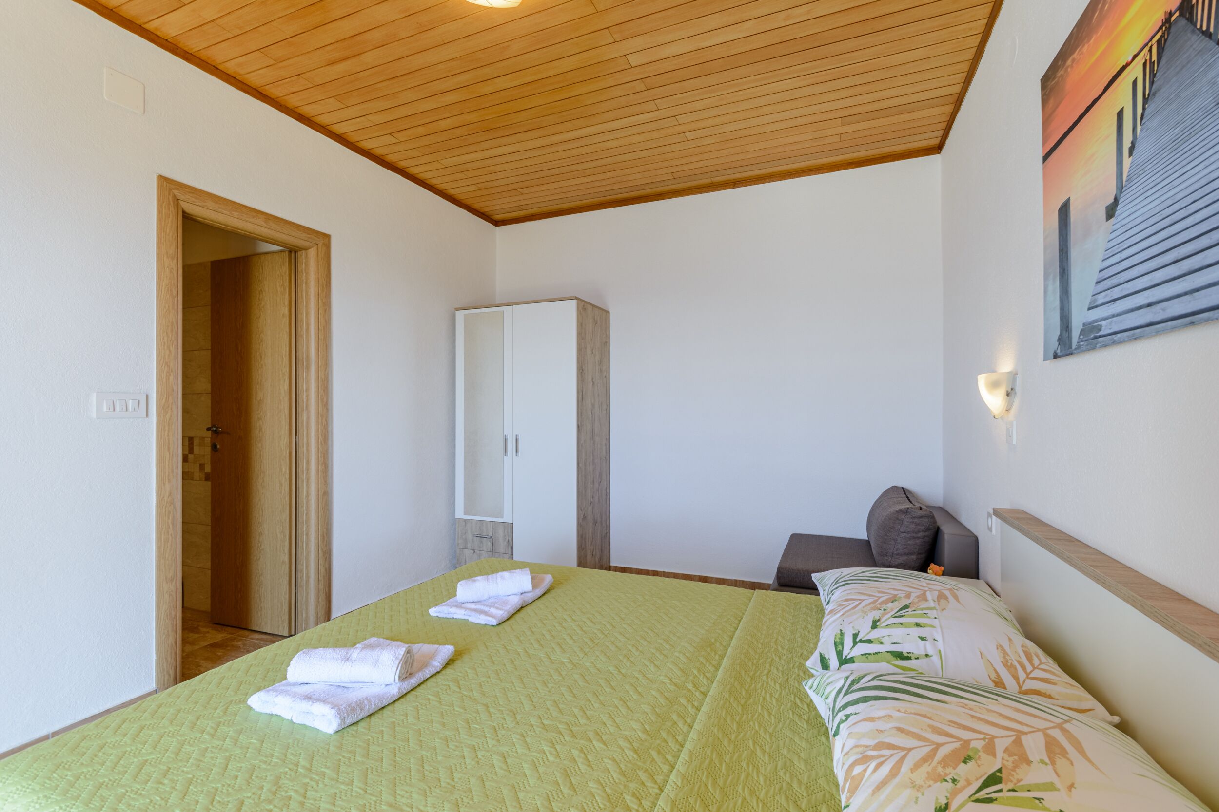A bedroom with a wooden ceiling, a bed with green bedding, a wardrobe, and a painting on the wall.