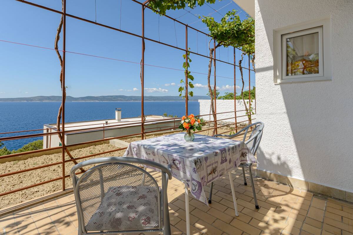 A terrace with a table and chairs, a view of the sea, and a white building with a window.