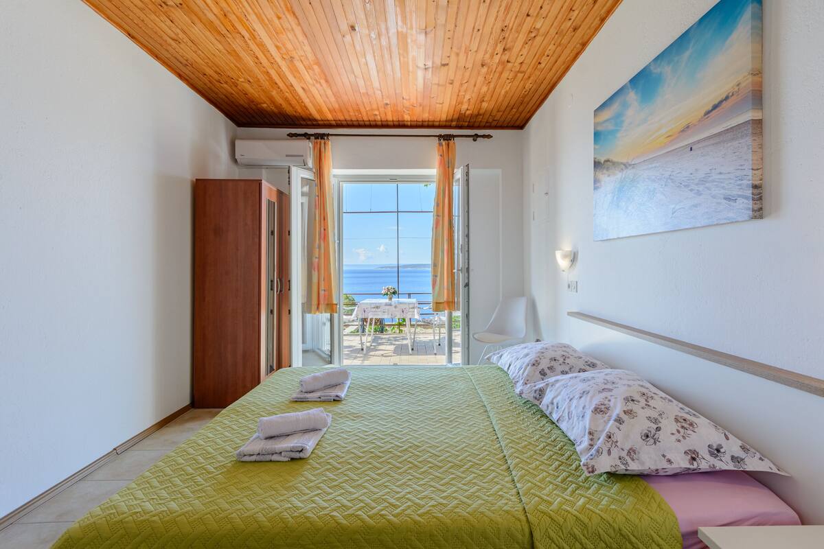 A bedroom with a bed, a wooden ceiling, a painting, and a view of the sea.