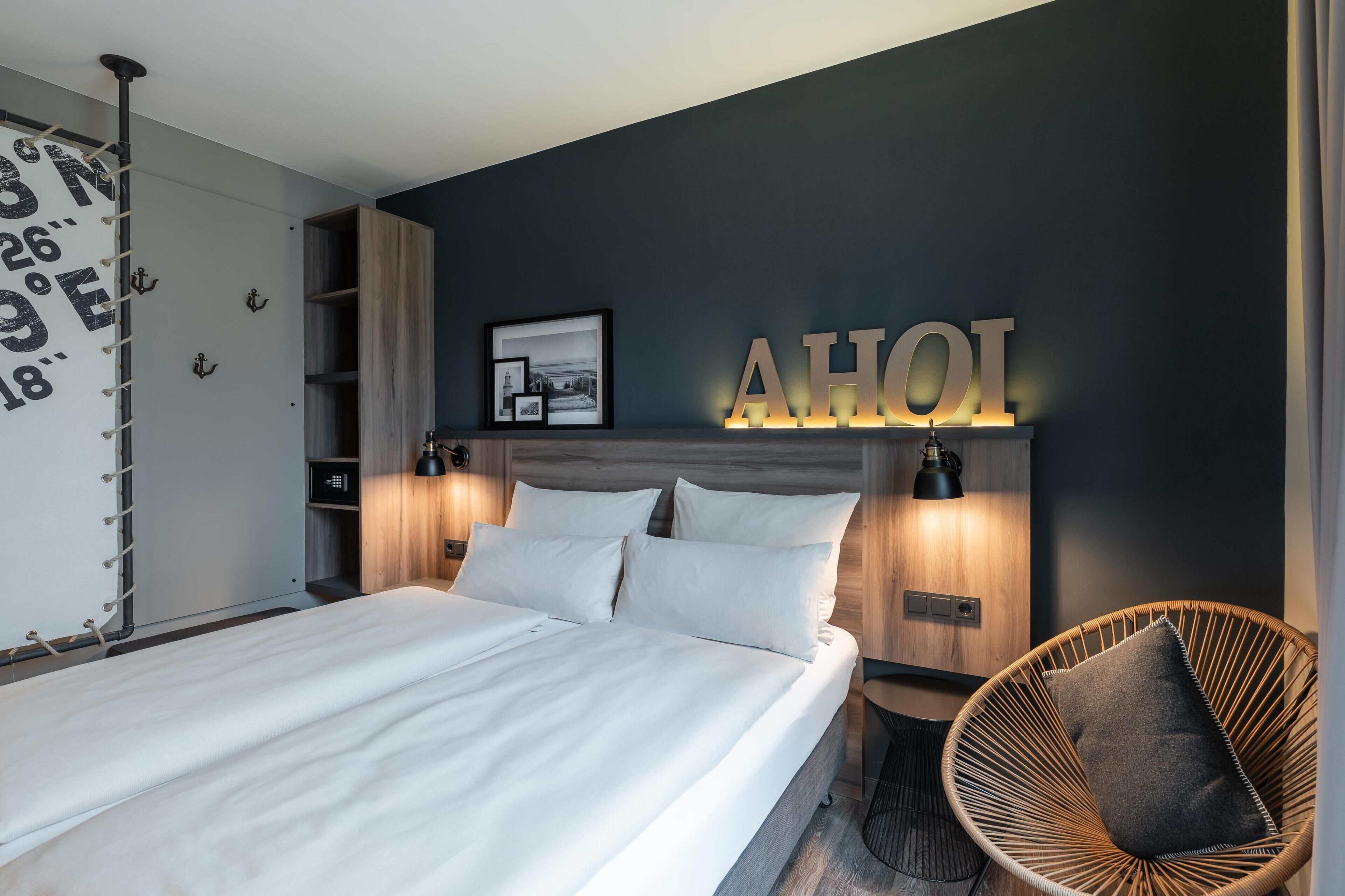 A modern bedroom with a large bed, a headboard with a 'AHOI' sign, a wicker chair, and a wall-mounted shelf.