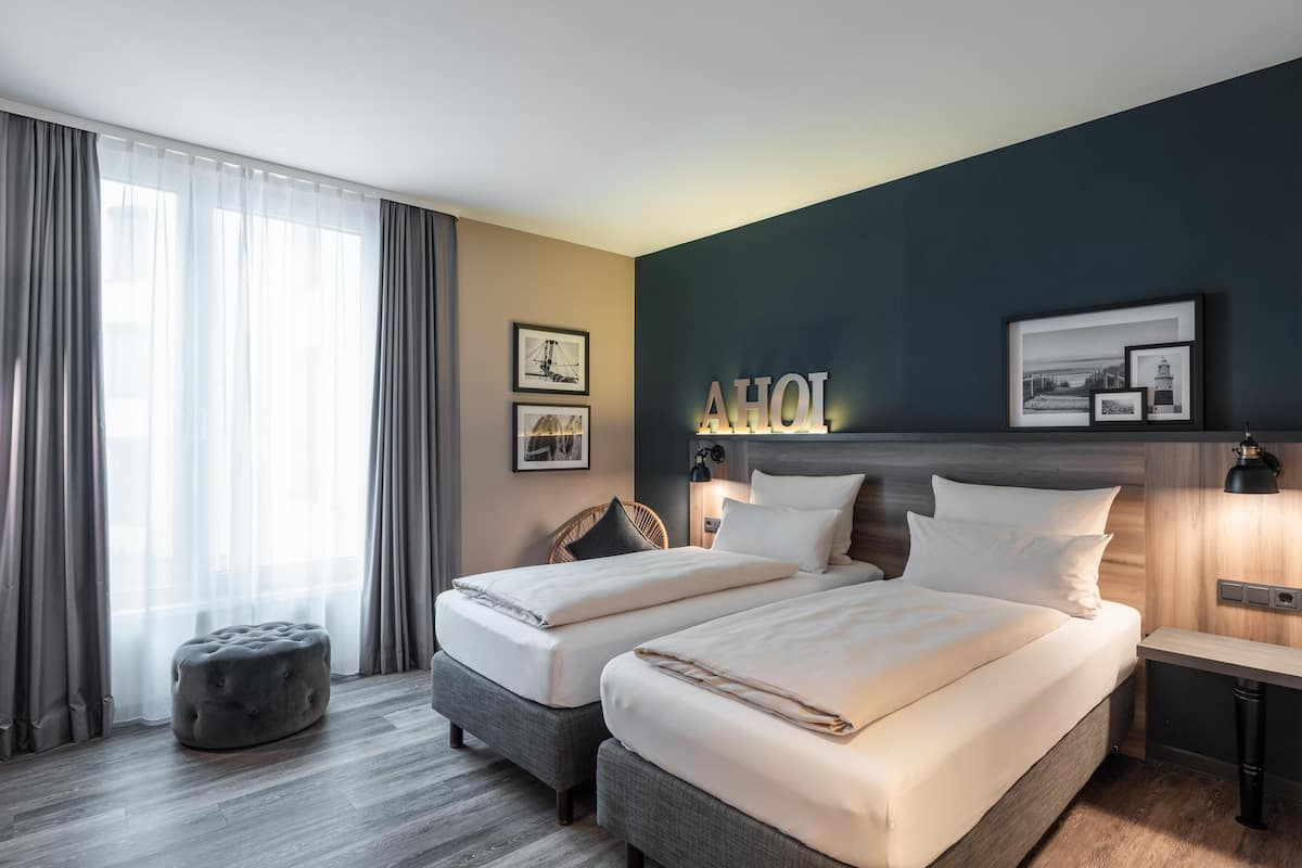 Hotelkamer met twee bedden, een grijze poef, een groot raam met dunne gordijnen en wanddecoratie.