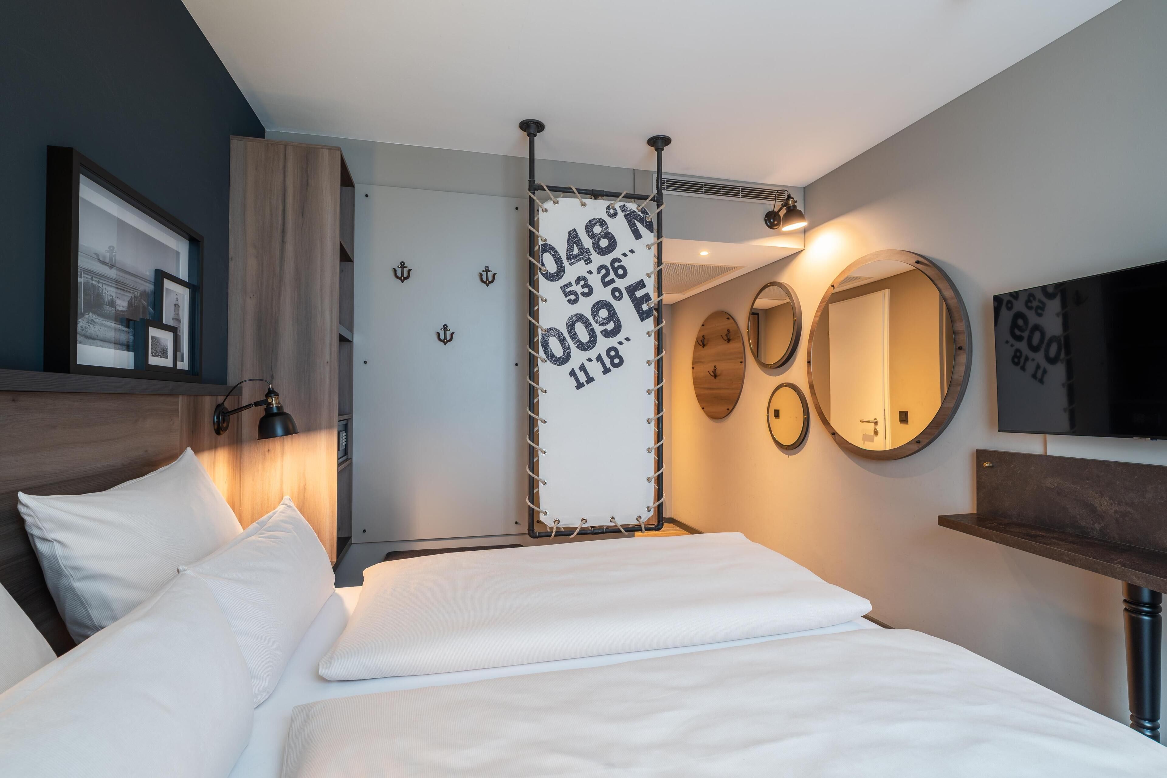A neatly made bed with white linens, a wall-mounted mirror, and a decorative sign with coordinates.