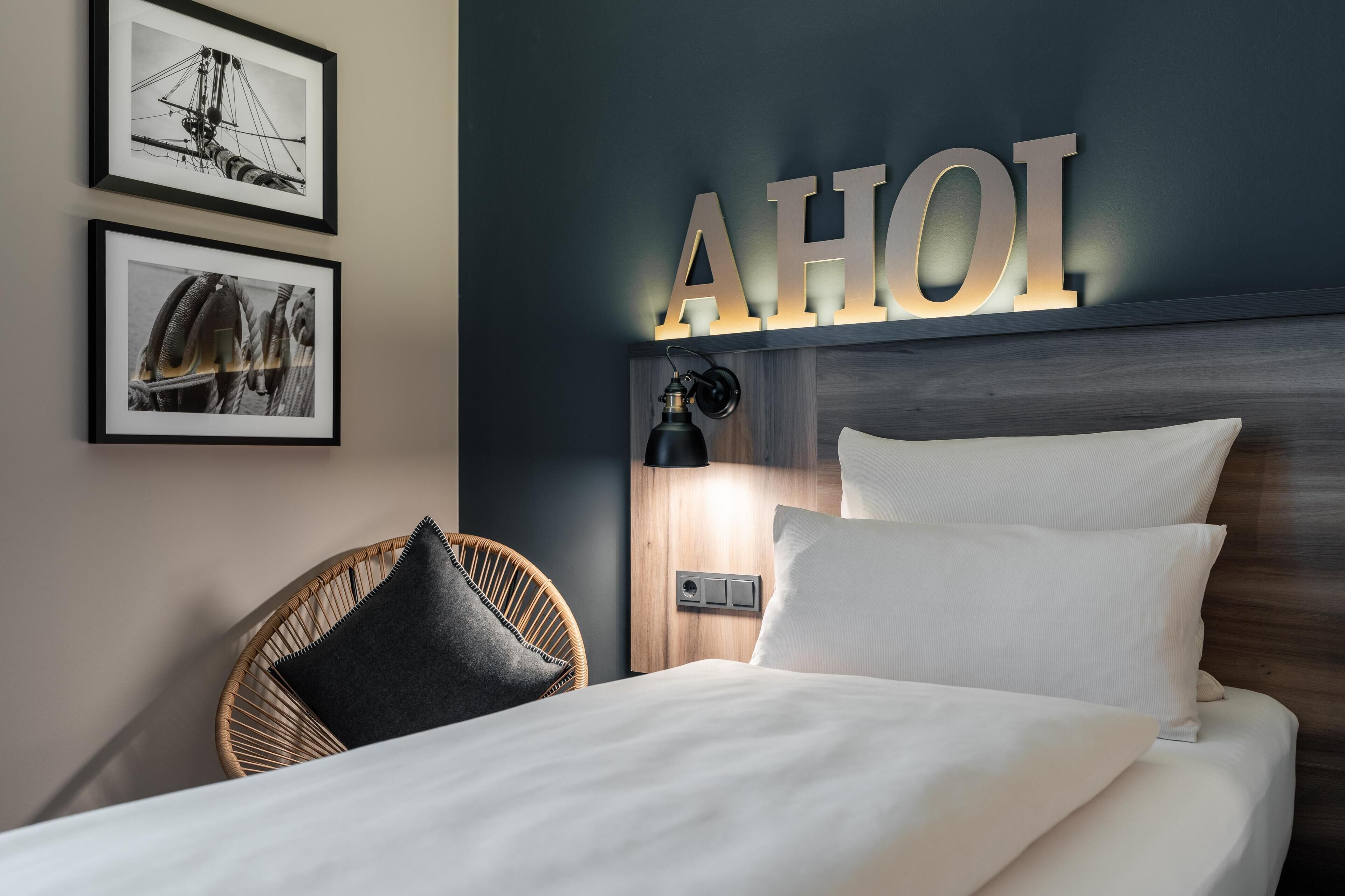 Una cama bien hecha con sábanas blancas, una silla de mimbre con cojín negro y una lámpara de pared.