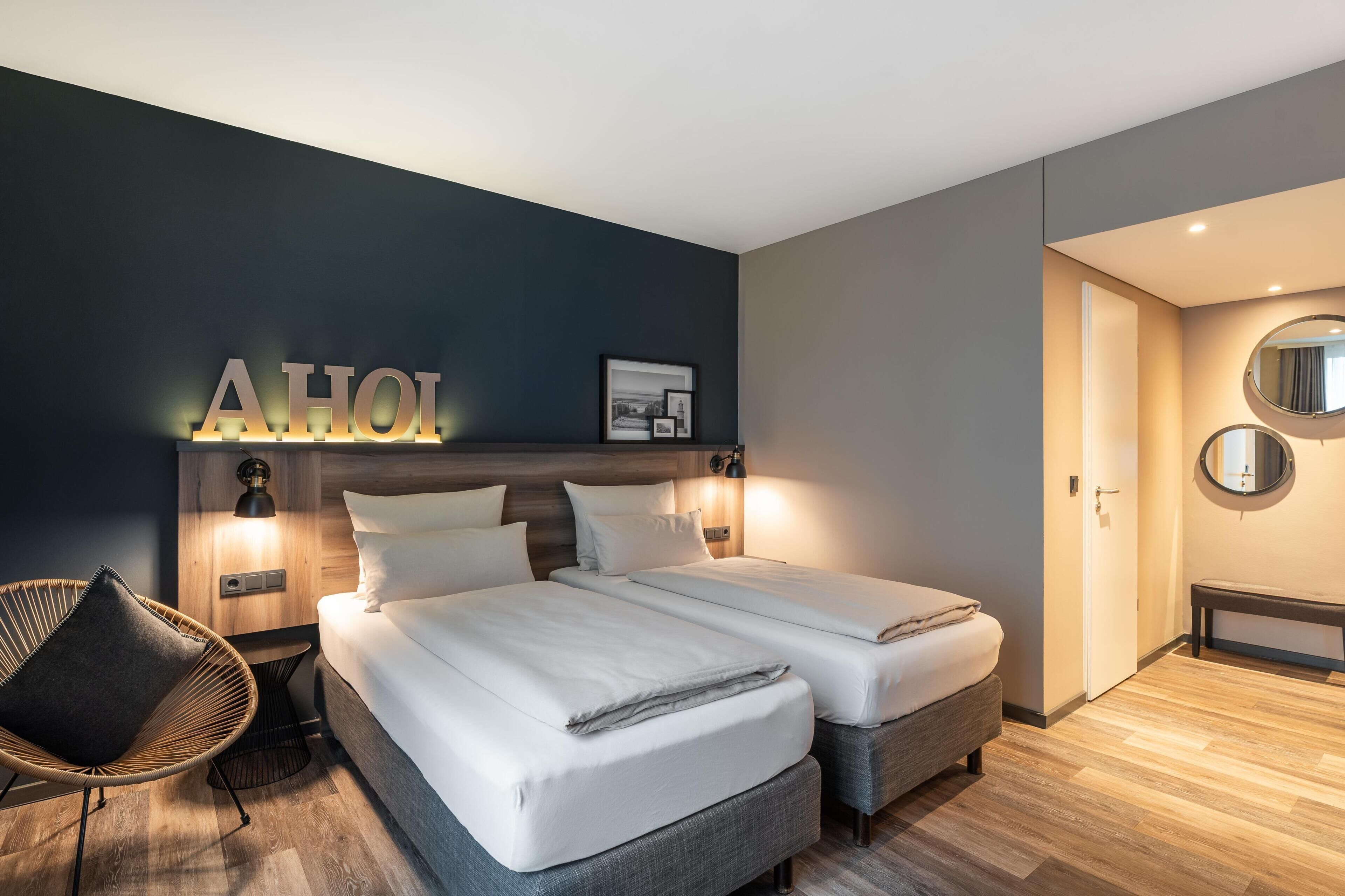 Una habitación de hotel moderna con dos camas, una lámpara fijada a la pared, un cuadro enmarcado y un banco.
