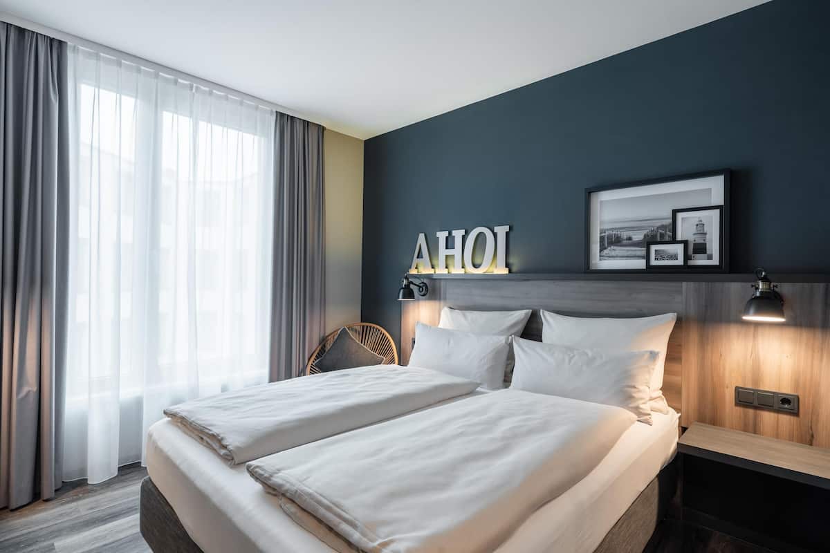 Een hotelkamer met een bed, een hoofdeinde met een lamp, een aan de muur hangend schilderij en een raam met gordijnen.