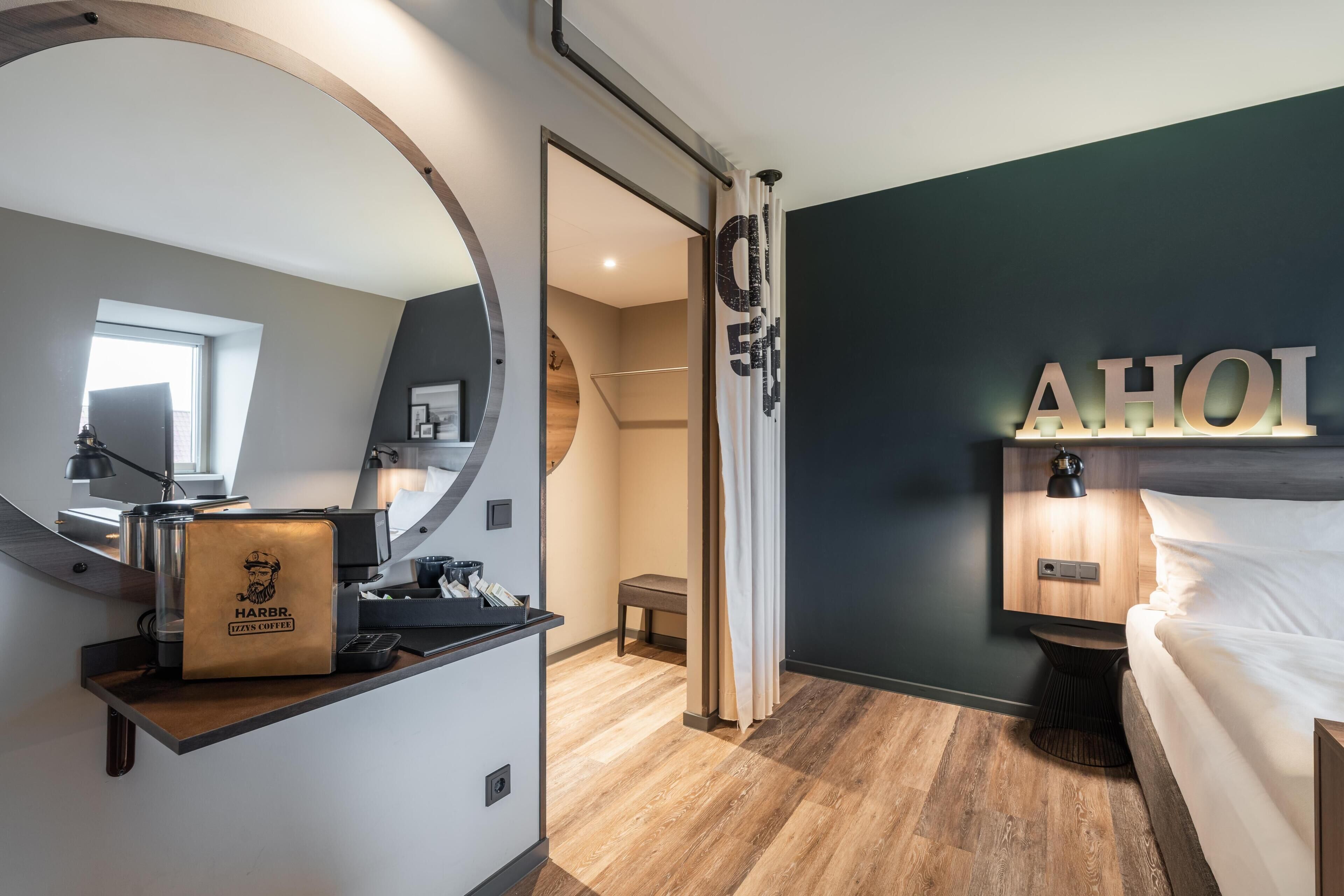 A modern hotel room with a bed, a desk with a coffee machine, a large mirror, and a wall-mounted light.