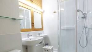 Kylpyhuoneessa on lasiseinällä varustettu suihku, kaksi pesuallasta, wc ja pieni valkoinen jakkara.