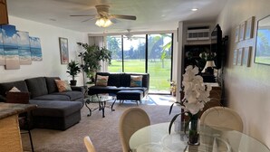 A spacious living room with a large sofa, a glass coffee table, and a dining area with a glass table and chairs. There is a ceiling fan, a large window with a view of a garden, and several potted plants. - Honu Kai@Turtle Bay- Pool, AC, Golf, Beach (Kahuku)