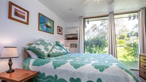 A bedroom with a bed, a bedside table, a lamp, a window with curtains, and a view of greenery outside. - Honu Kai@Turtle Bay- Pool, AC, Golf, Beach (Kahuku)