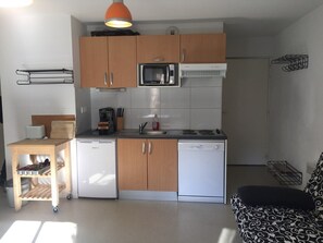 Una cocina funcional con armarios de madera, microondas, estufa y lavavajillas. Hay una pequeña mesa de comedor con una silla, y un sofá en la sala.