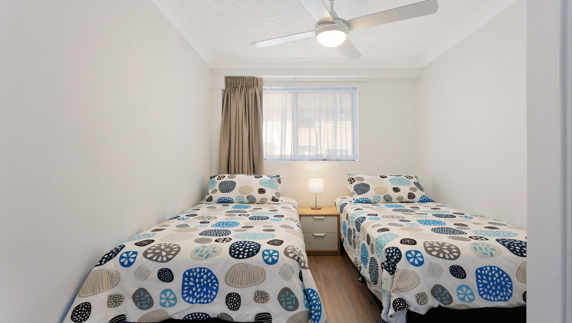 Une petite chambre propre, avec deux lits, un ventilateur de plafond, une fenêtre avec des rideaux et une table de chevet avec une lampe.