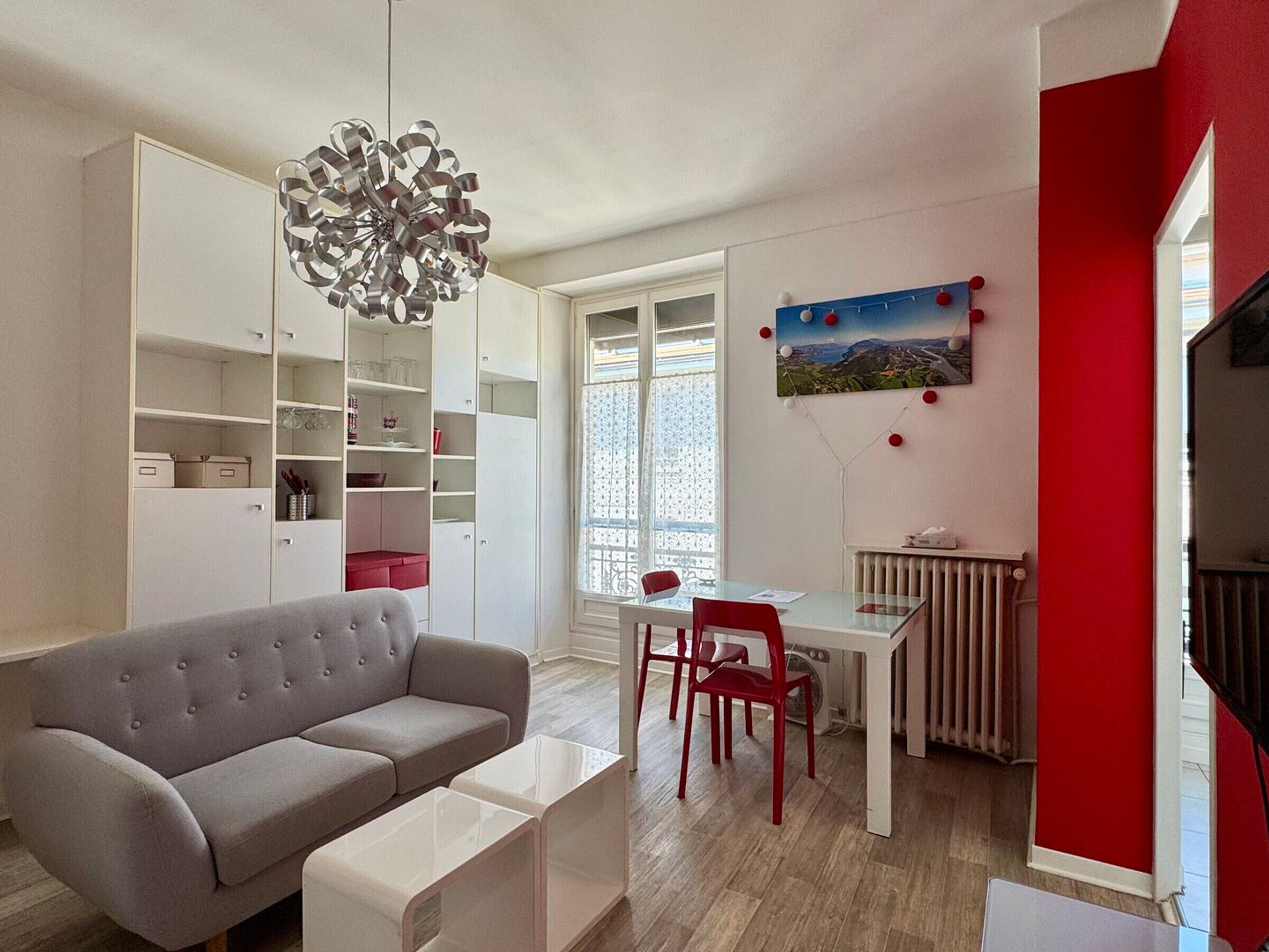 Un salon moderne avec un canapé gris, une table à manger entourée de chaises rouges, un meuble blanc et un lustre original.