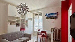 A modern living room with a grey sofa, a dining table with red chairs, a white cabinet, and a unique chandelier.