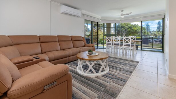 Ein modernes Wohnzimmer mit einer braunen Ledersofa, einem kleinen Couchtisch und einem Essbereich mit weißen Stühlen und einer Glastür zum Balkon.