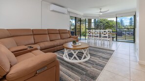 A modern living room with a brown leather sofa, a small coffee table, and a dining area with white chairs and a glass door leading to a balcony. - Parkside Marina 1 - CBD Location (Forster)