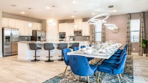 A modern kitchen with stainless steel appliances, a central island with bar stools, and a dining area with blue chairs and a glass table set for six.