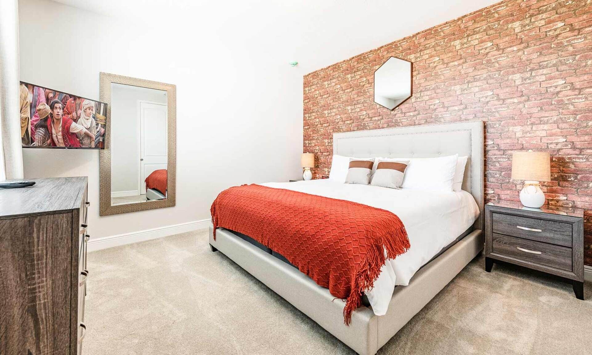A hotel room with a brick wall, a large bed with white bedding and a red throw, a nightstand with a lamp, and a mirror on the wall.