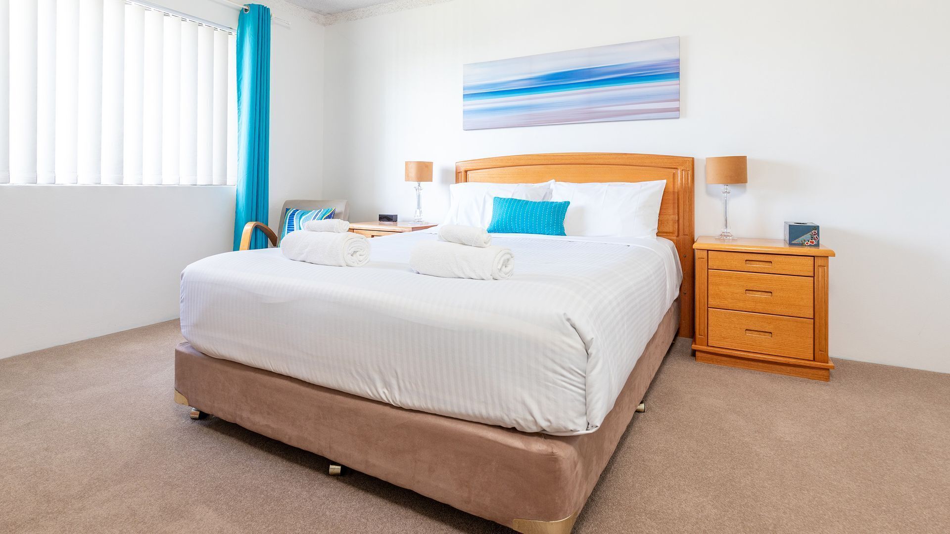 A neatly made bed with white linens, a wooden headboard, and a blue pillow. There is a wooden nightstand with a lamp and a small box on it.