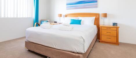 A neatly made bed with white linens, a wooden headboard, and a blue pillow. There is a wooden nightstand with a lamp and a small box on it.