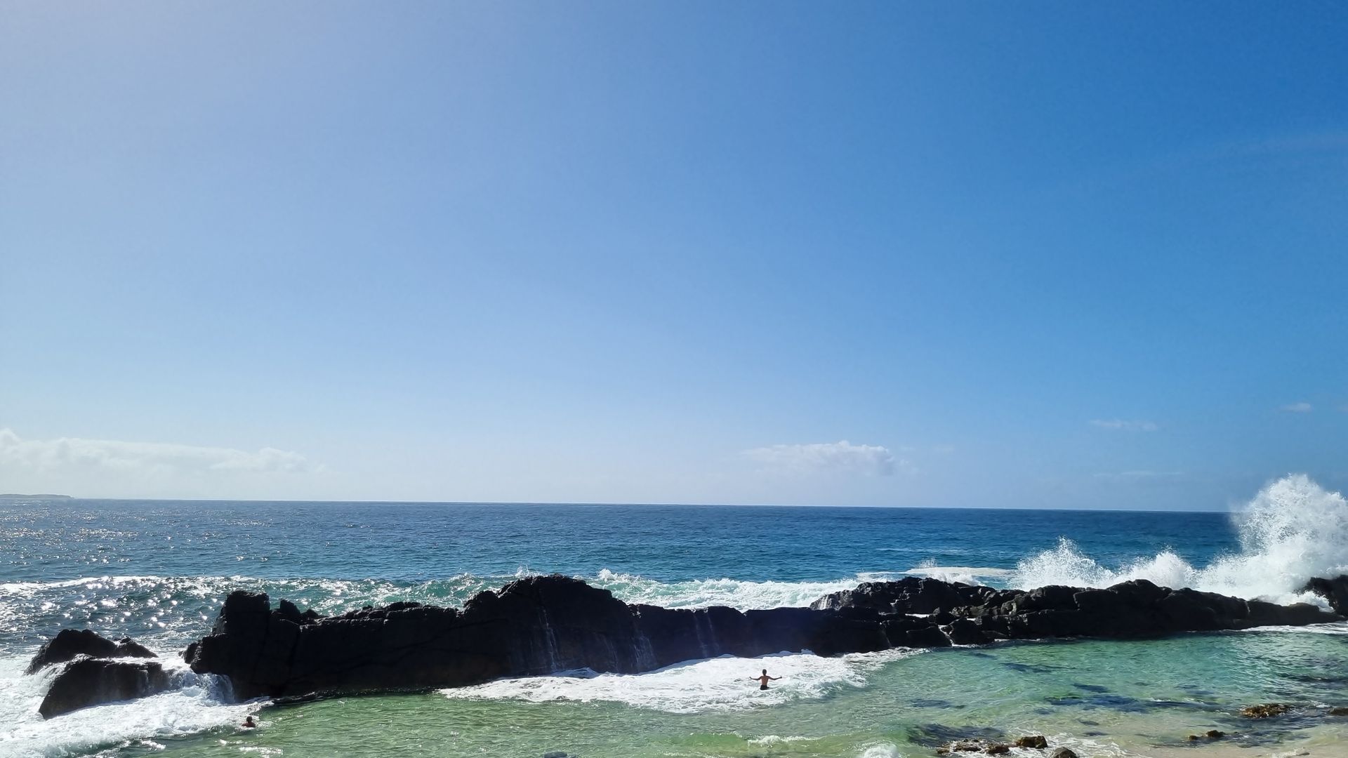 A coastal scene with clear blue water, waves crashing against dark rocks, and a person swimming in the water.